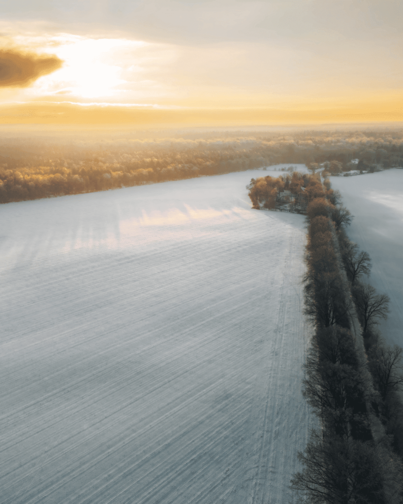 Drohnenaufnahme einer verschneiten Straße im Winter, eingefangen bei Sonnenuntergang, mit einer malerischen Landschaft und goldenen Lichtreflexionen im Schnee.