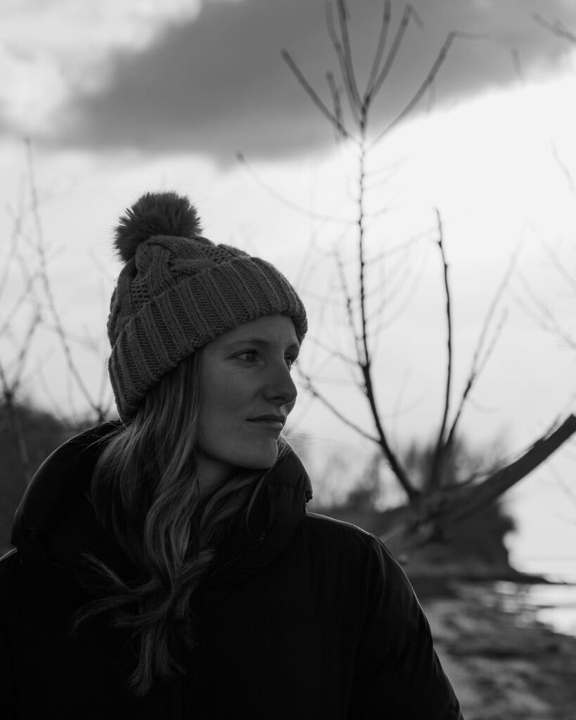 Portrait einer Frau am winterlichen Strand der Ostsee. Sie steht im kühlen Wind, umgeben von karger Natur und einer rauen, grauen See. Die Szene strahlt Ruhe und Melancholie aus, während die kühle Winterluft die Stimmung der Ostsee perfekt einfängt.